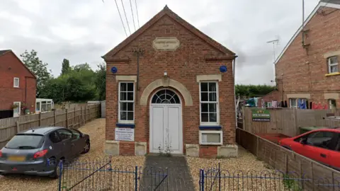A blue gate leads on to a short pavement which goes to a Methodist church. The church has a white door and two white windows. There is a sign above the entrance. 
