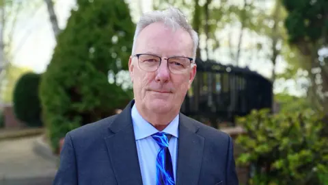 Nesbitt is wearing a navy suit with a blue shirt and a blue tiw. He wears glasses. Trees and bushes are behind him. 