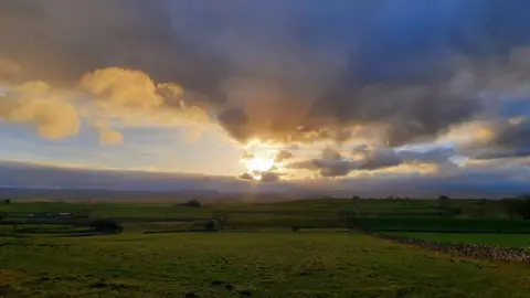 Weather Watchers/Gadabout Dark clouds are gathered at the top of a blue sky. The sun is bursting through and rays of light are shining onto the field below. The field that stretches to the horizon is green with a dry stone wall running across it.