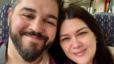 Handout Chris Sherwin, who has beard and black hair, and Amy Hawkswood, who has long dark hair, sitting together on a train carriage, smiling. The pattern of the seat fabric is behind them.