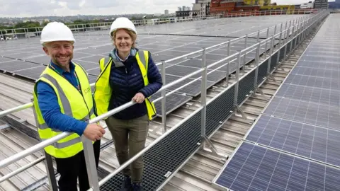 two people in high vis jackets stand on an elevated metal walkway on top of a roof with solar panels 