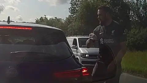 A man with short dark hair and beard looks on at a car angrily on a country road