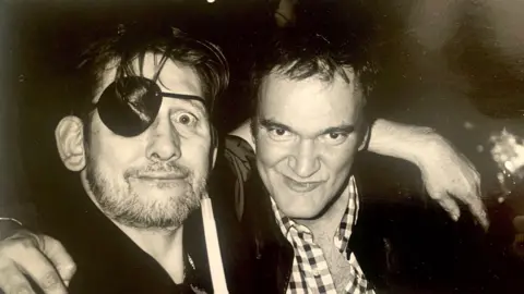 Johnnie Shand Kydd A black and white photograph of Shane MacGowan and film director Quentin Tarantino. Shane is wearing an eye patch and has his arm around the shoulder of Quentin - who in turn has his arm around Shane's shoulder. They are both looking into the camera. 