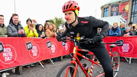 A man in black cycling gear, a red helmet and a red nose is leaning forward on a red tandem bike. There are red barriers in the street which read "Comic Relief Red Nose Day" with people stood behind them taking photographs on their phones.