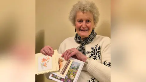 An older woman, wearing a white jumper, holds up colourful homemade cards and smiles.