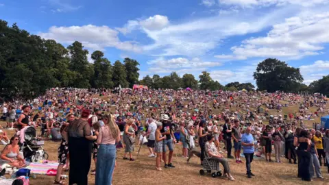 Ollie Ward/BBC Hundreds of people enjoying a festival within a park on a sunny summer's day. 