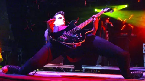 Sam Endicott of the Bravery does a partial splits as he plays a black bass guitar. He is dressed mainly in black and his a black quiff.