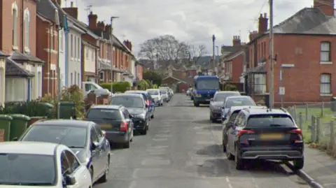 Google A residential street with terraced houses on either side and vehicles parked on both sides of the road