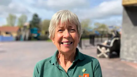 The image shows a woman standing in front of a ride at a Devon theme park. She is wearing a green t-shirt with the Visit Devon logo. 