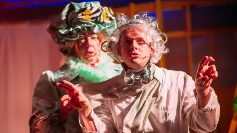 Two male actors on stage. The one in front is wearing a beige shirt, scarf and hat. He has unruly white, curly, hair and has large goggles around his neck. He is gesticulating with his hands. The man behind him is dressed as a  pantomime dame. He is wearing a dress and has green feathers around his neck and a green bonnet on his head featuring yellow pound signs.