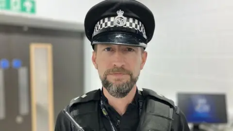 BBC A police officer in a black and white chequered hat with a crest on it. He is wearing a black vest, radio and lanyard.