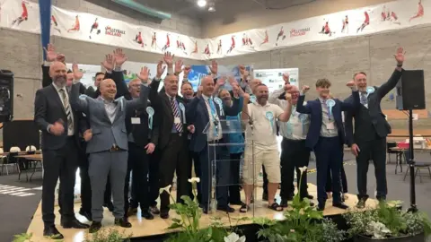 Laura Coffey/BBC A group of men and a woman standing on a stage in a sports hall. Most are dressed in suits some with ties. They are smiling and cheering. Many are wearing white and blue rosettes.