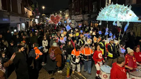 Participants in the Bedminster Winter Lantern Parade walk down East Street. The photograph is taken at night and hundreds of people are visible, many of them carrying colourful, illuminated lanterns.