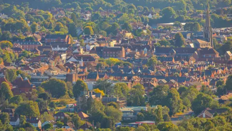 Getty Images View of Dorking town center from the Surrey Hills. The sun shines on the buildings, between the green trees there are many houses and an obvious church tower. 