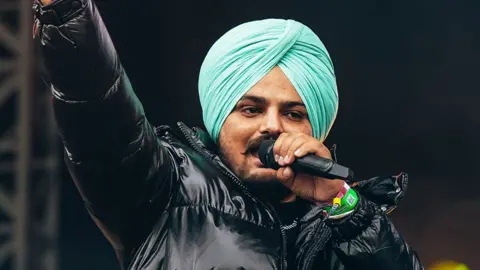 Getty Images A man wearing a black jacket, performing on stage singing into a black microphone, wearing a light blue turban (religious head covering for Sikh's).