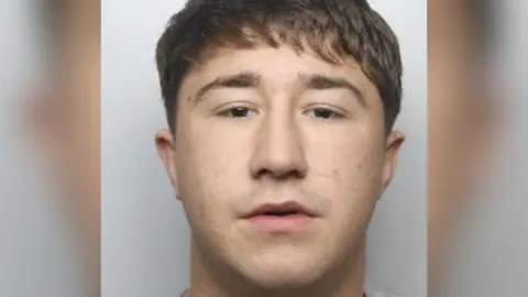 South Yorkshire Police A man in his early 20s stares at the camera in a police custody image. He has brown hair and is wearing a grey top.
