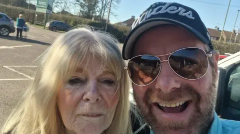 Hudgell Solicitors Nicholas Gibbs, with his mother, who has long blonde hair. They are in a car park, he has on a blue cap and sunglasses and has a beard. They are both looking at the camera. 