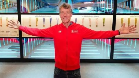 Chris Finill Chris Finill, wearing a red tracksuit top, grins as he stands with arms outstretched in front of a display case showing his London Marathon medals.