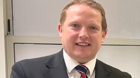 Manx Care A headshot of a man with short, light brown hair and blue eyes wearing a grey suit with a blue and red striped tie. 