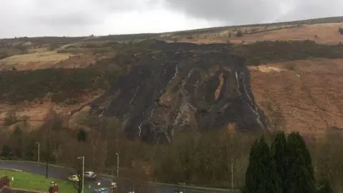 A coal tip has slipped leaving a blackened hillside. Either side of the landslip the grass and bracken is orange and yellow in colour. There is a grey, cloudy sky and in the foreground there is a road with lamp posts and trees either side of it and a couple of chimney stacks are visible in the bottom left hand corner.