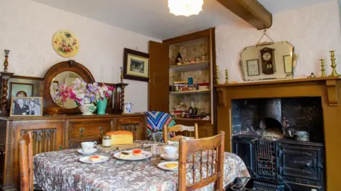 North Kesteven District Council An interior view of the museum. There is a table with a floral cover on it and various items of old-fashioned furniture and a stove set within a wooden fire surround. There is a beam on the ceiling and photographs on the wall.