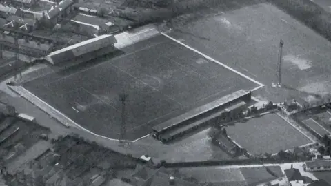 Aero Pictorial Limited A black and white aerial shot of Cambridge City's Milton Road ground in 1960. The ground takes up much of the shot and shows two covered stands at its top and bottom sides. Tall scaffolding towers for spot lights can be seen at all corners of the ground. On the top left and along the bottom can be seen houses and gardens 