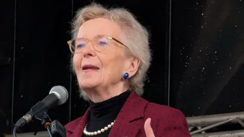 Robinson has short blonde hair and is wearing glasses, blue earrings, a pearl necklace, a black top and red blazer.