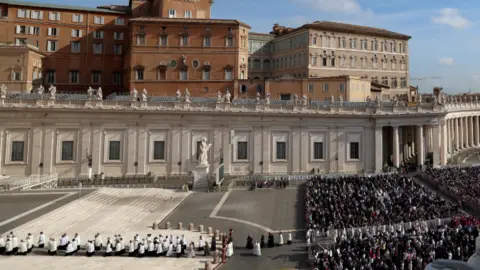 The faithful, on the right, gather normal clothes in the Plaza de San Pedro while the members of the clergy, on the left, gather white. There are statues and large buildings on the crowd.