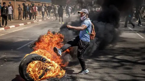 AFP via Getty Images A protester, in a light blue jersey and a cap, kicks a burning tire amid clashes with Malagasy security forces during a rally calling for constitutional reforms in Antananarivo