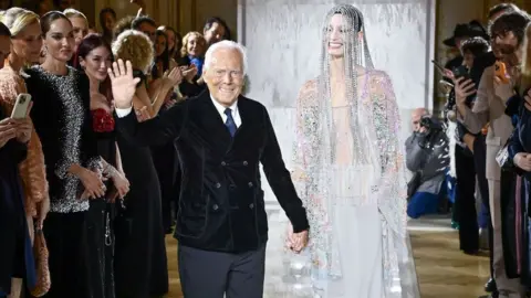 Getty Images An elderly man in a velvet jacket holds the hand of a tall model, wearing beads across her face and a sheer, embellished dress.