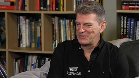 Graeme Lowdon is sitting on a grey chair side on and smiling. He is wearing a black shirt with the Cadillac logo on it in white. Behind him is a book shelf separated into compartments with lots of books in them.