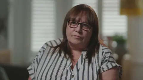 A woman with dark hair and thick rimmed glasses is sat in front of a blurred out living room, which has a plant and shutter blinds in the background. She is wearing a cream and black pin-stripe shirt and looks towards the camera.