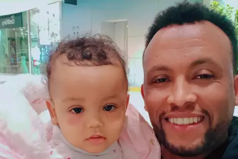 Family photo Nestere and his young daughter look directly at the camera smiling. The little girl is wearing a pink coat.