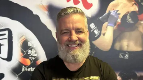 BBC Danny Corr has grey hair which is slicked back and a grey beard. He is smiling at the camera and wearing a black t-shirt with a camouflaged logo.