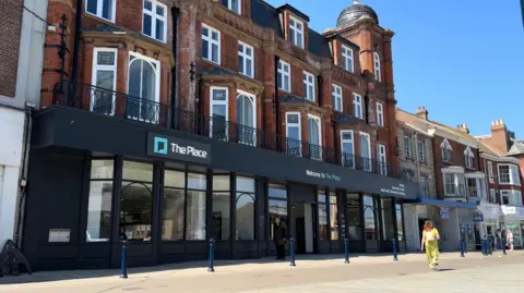 Andrew Turner/BBC A large red-bricked building with a dark grey frontage on its lower floor is in view as a woman wearing a yellow outfit walks past in Great Yarmouth's Market Place