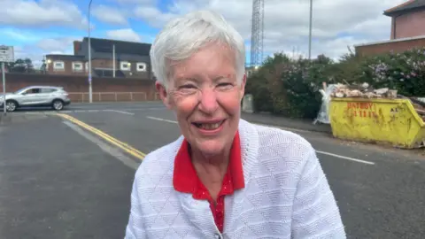 An older woman, with short grey hair, wearing a red t-shirt and a white cardigan. She is standing on a path with a road behind her. There is a yellow skip behind her left shoulder. 