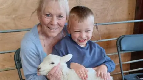 A smiling woman with light blonde hair, wearing a grey cardigan and sat on a black fold-out chair with a young smiling blonde boy wearing a blue jumper on her lap. On his lap is a white baby lamb, and both the woman and boy have their hands on the lamb.