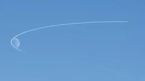 Donald Milne Blue sky and plane trail making an arc around the moon.