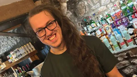 A man in black rimmed glasses, with long dark wavy hair swept forward over one shoulder. He is in front of a pub bar and is smiling.