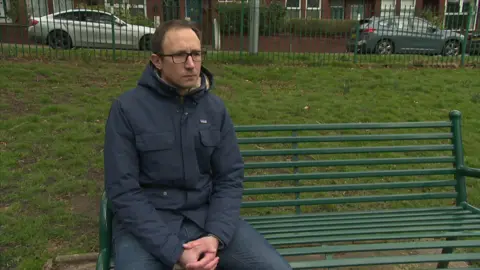 A dark-haired man sits on a green metal park bench, wearing glasses, with his hands clasped on his knees 