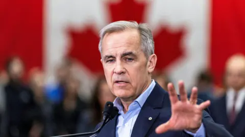 Reuters Canadian Prime Minister Mark Carney, wearing a dark blue suit and light blue open-necked shirt, gestures with his left hand during a speech