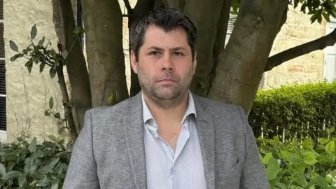 Leigh Frost looks serious as he stands in a garden with a tree behind him. He has dark hair and brown eyes and is wearing a grey jacket and a grey and white striped shirt. 
