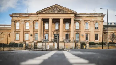 The exterior of Dundee Sheriff Court. The photo is taken from ground level so traffic lines are visible. It is a large sandstone building with four columns at the front.