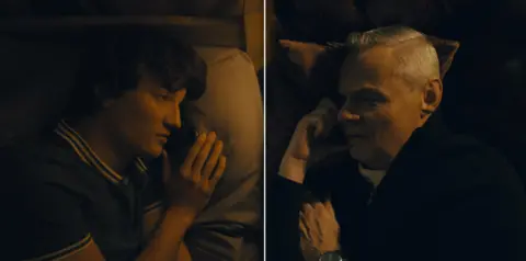 5 Broadcasting Limited / Paramount / Wonderhood Studios/ PA Split screen with Morgan and Clunes. They are facing each other while lying on a bed, but not in the same place. Morgan wears a green t-shirt with stripped collar, while Clunes wears a black quarter zip jumper. They hold a phone to their ear.