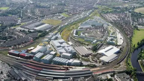 Homes England/Network Rail The photograph is an aerial view of a large urban area featuring a mix of industrial, residential, and green spaces, with a network of roads, railways, and buildings. At the bottom of the image there are multiple parallel train tracks and platforms. Several long trains are visible, some with red roofs.
Surrounding the rail area are large industrial buildings with flat roofs, mostly white or light grey in colour. 