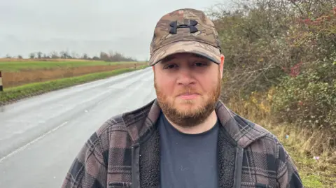 John Devine/BBC James Fuller, has a reddish beard and moustache, his head is covered with a camouflage baseball cap, he is wearing a heavy, tartan fleece jacket with a black t-shirt underneath. Behind him is a Fen road positioned next to a drain (like a river) the water is not visible, but it just over a grass verge about 30cm (1ft) high.