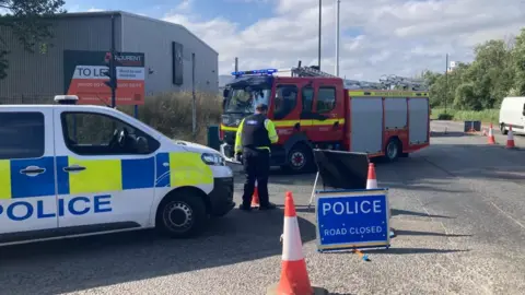 A police van and fire engine on a road with a road closed sign and cones.