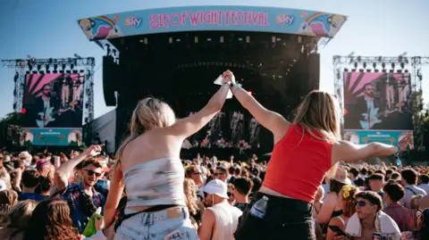 Isle of Wight Festival - Day Rutherford View of Isle of Wight stage from the crowd. The photographer is standing immediately behind two women who are sitting side-by-side on the shoulders of two men.