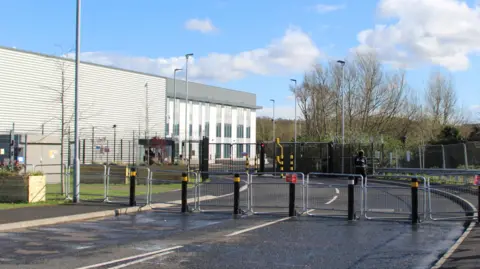 The Elbit Systems UK factory in Filton, near Bristol. There are metal gates blocking access to the building, which is white and grey. 