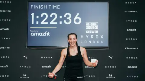 Beth Littlewood A woman wearing all black sports kit and her brown hair in two plaits. She is holding a small card in one hand with the words Hyrox London on it. The other hand is clenched in a fist. She is smiling and looking at the camera. Behind her is a board with the Hyrox London logo in one corner and then the words: "FINISH TIME: 1:22:36". Under that it reads "presented by amazefit". 